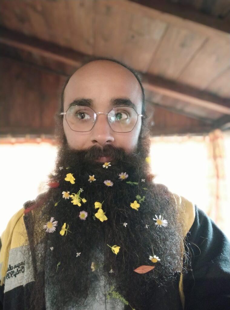There shall be flowers of blessings... A profile photo of mine taken in 2020. I have a long beard, balding head, and I wear oddly shaped hexagonal glasses. There are lots of flowers in my beard, that my friends decorated me with. I am facing the camera but looking away. In the blurry background you can make out that I am on the porch of a wooden cabin.